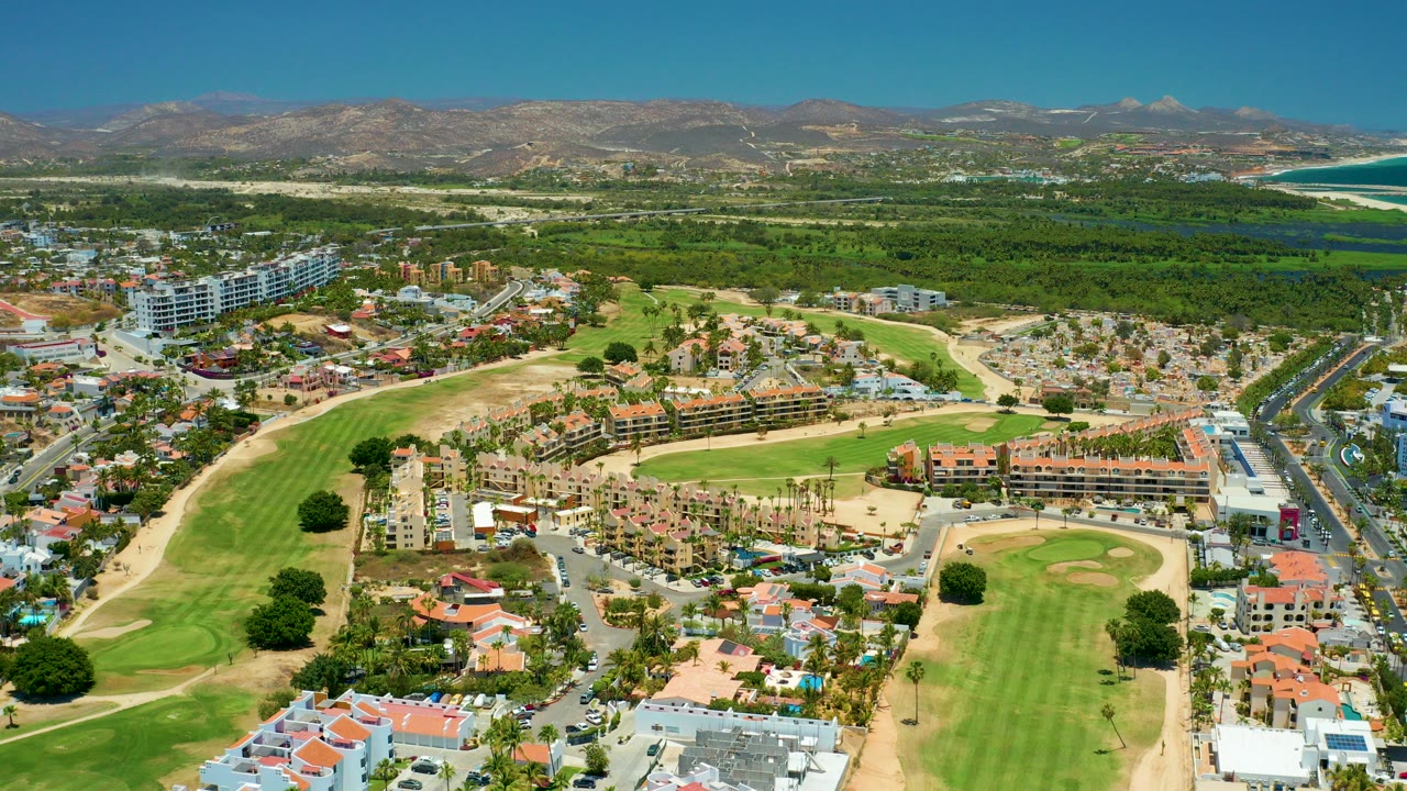 Wide aerial frame over San Jose del Cabo with green golf fairways in the foreground, residential buildings and roads in the center, mountains on the horizon, distant ocean at right, and no visible people.