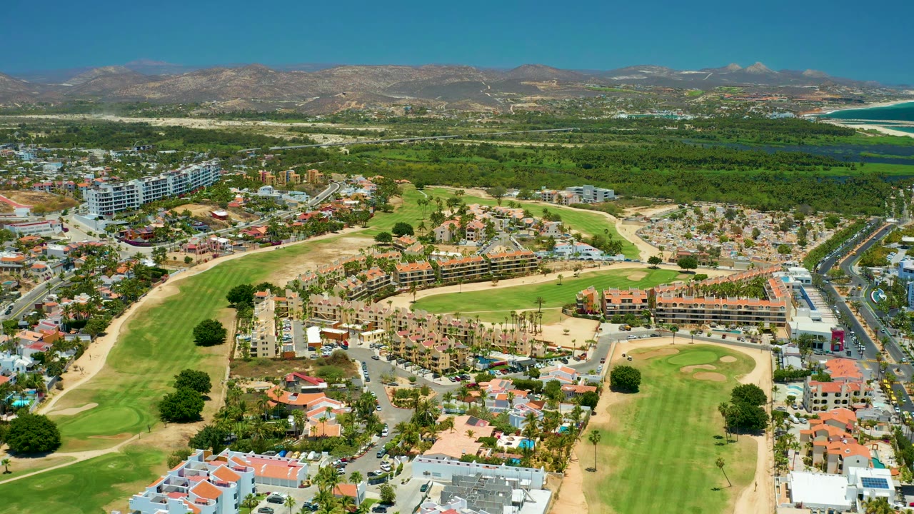 Aerial view in San Jose del Cabo showing the golf course shifted across the lower frame, clustered residences beside a road, green vegetation beyond, mountains in back, distant ocean at upper right, and no visible people.