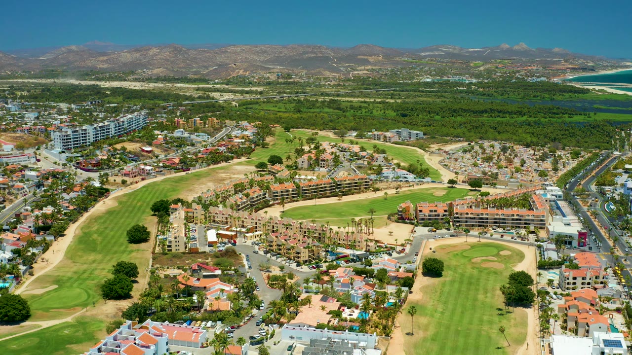 San Jose del Cabo aerial frame with apartment-style buildings at left, fairways and sand traps below, a straight road through the right side, broad greenery behind the neighborhood, mountains beyond, and no visible people.
