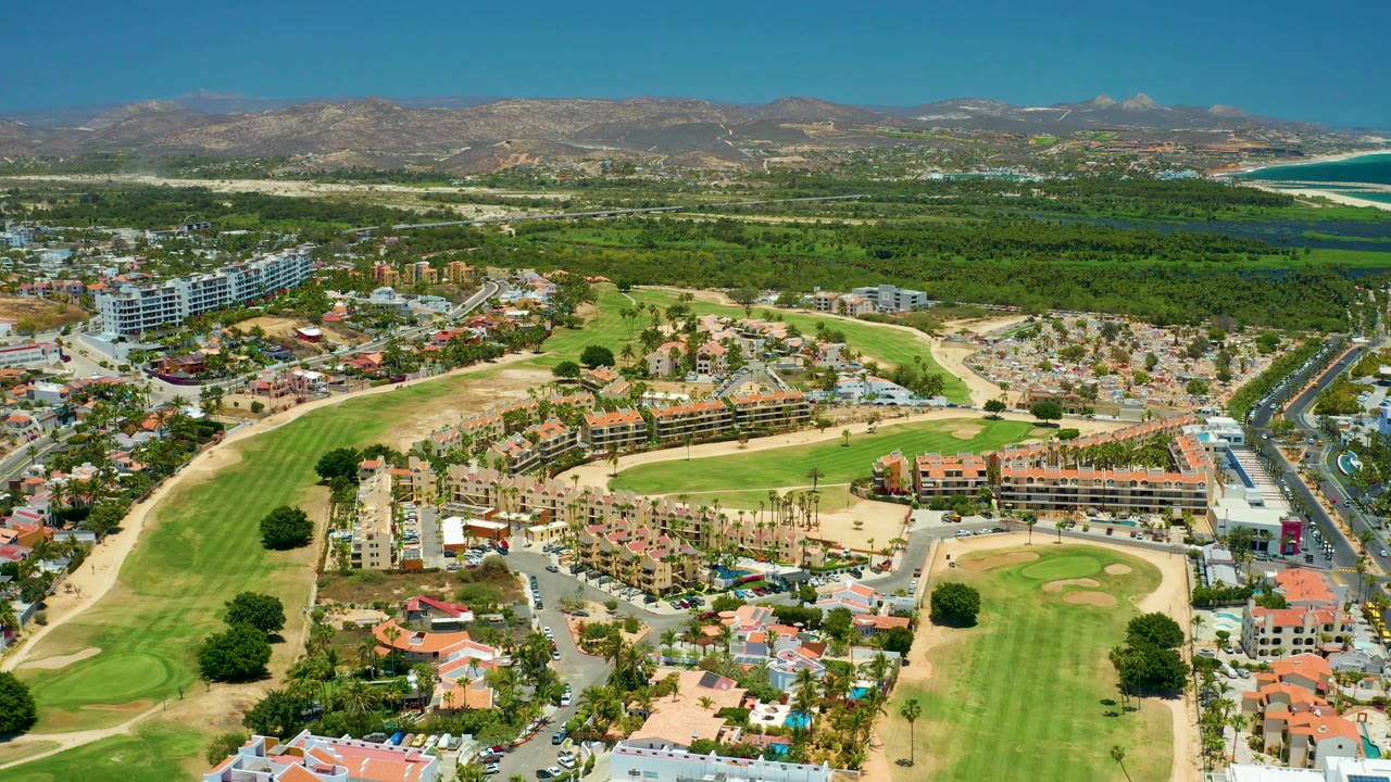 Aerial panorama of green fairways, tan bunkers, homes, road, mountains, and coastline.