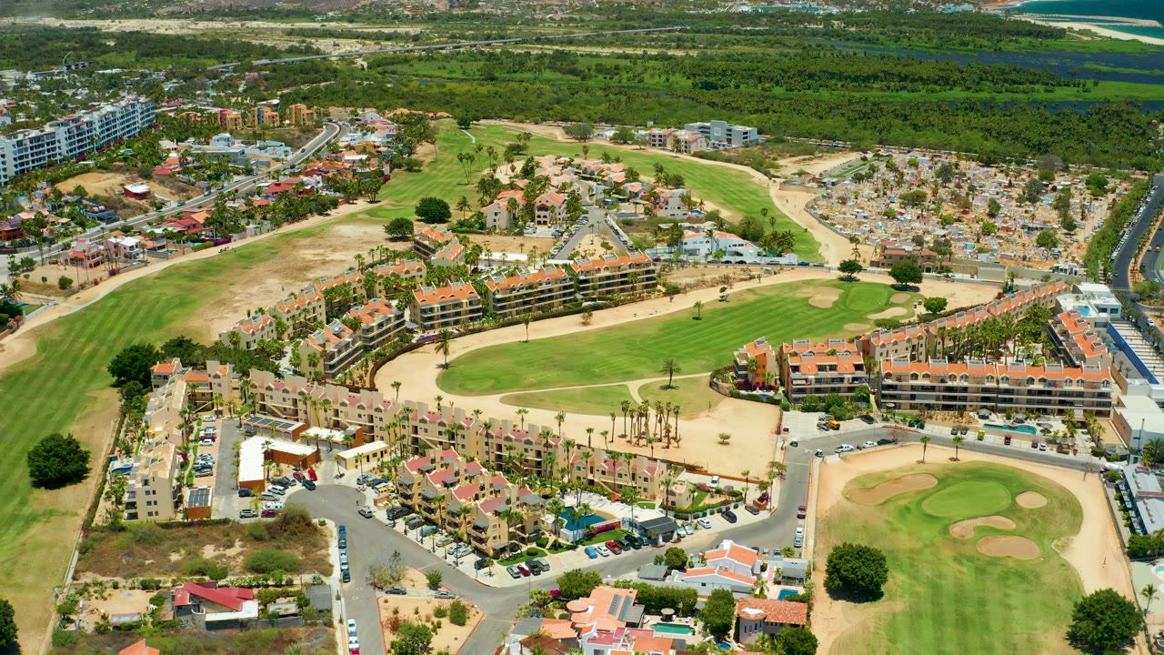 Aerial frame over San Jose Del Cabo showing red-tile residential buildings, parking areas, curved roads, golf greens, sand traps, and no visible people.