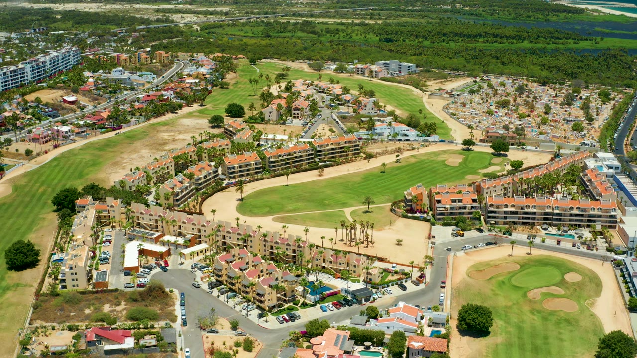 San Jose Del Cabo aerial frame with clustered residential roofs in the foreground, a road network, golf course grass and bunkers, parking areas, and no visible people.