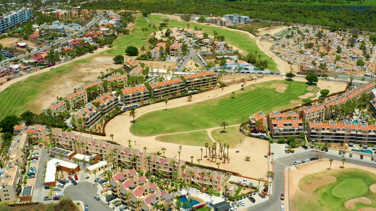 Opening aerial frame over San Jose Del Cabo with orange-roof buildings, green fairways, sand traps, roads, parked vehicles, and no visible people.