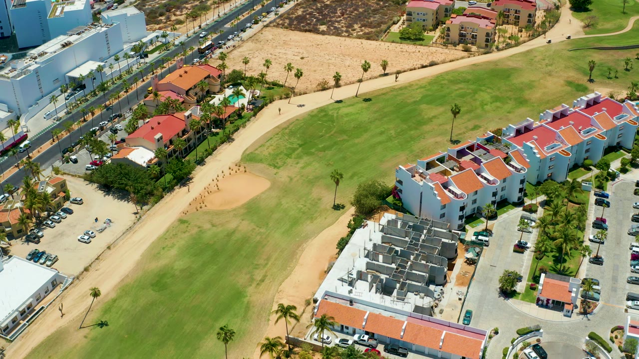 Aerial frame in San Jose Del Cabo with the sand trap left of center, a broad green fairway, white and red-roofed buildings, parking rows, cars on a road, and no visible people.