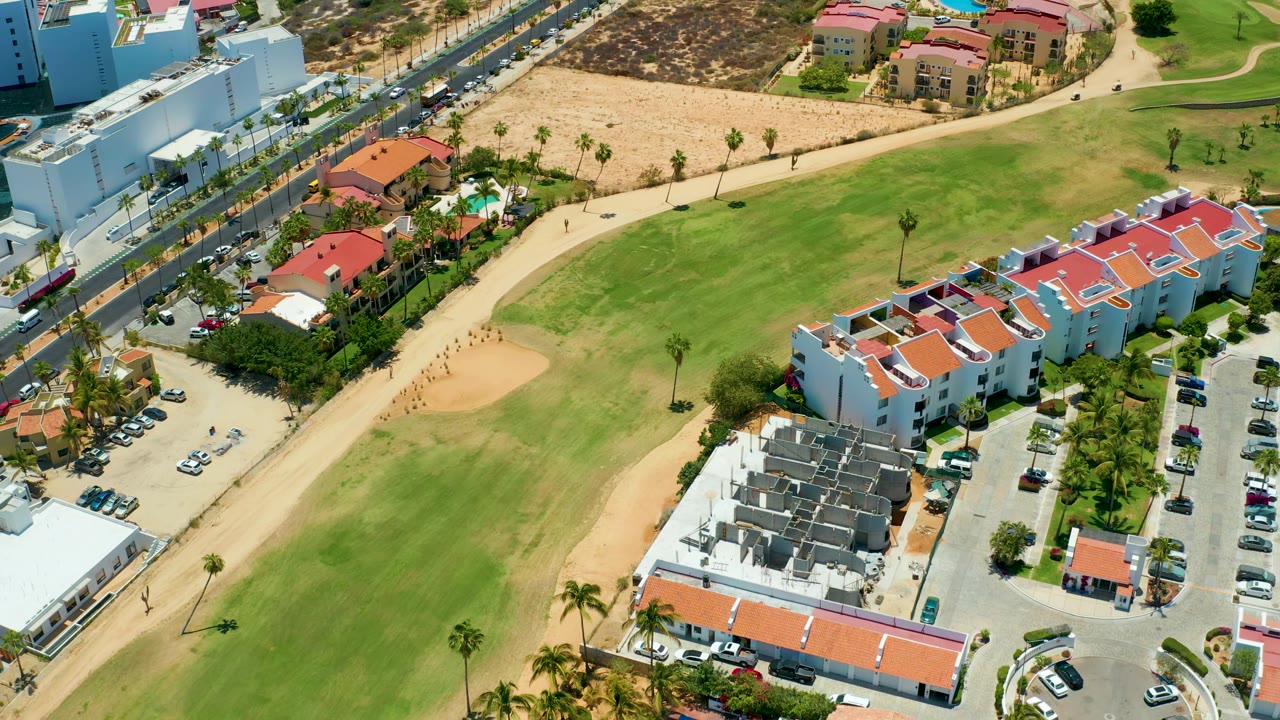 Drone-view frame over San Jose Del Cabo showing the fairway curving past red-roofed residences, a tan sand area, a lined road, parking lots, palm trees, and no visible people.