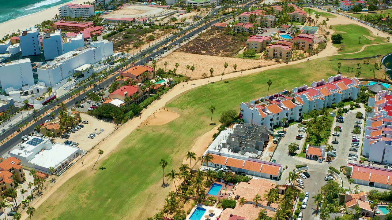 San Jose Del Cabo aerial frame with the beach and surf along the upper edge, a busy coastal road through the center, golf course grass in the foreground, white buildings, red rooflines, parked cars, palms, and no visible people.