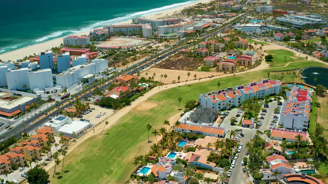 Aerial view in San Jose Del Cabo, Mexico, showing the beach and ocean along the upper left, a coastal road, red-roof homes, green lawns, white buildings, and a dark pond at right, with no visible people.