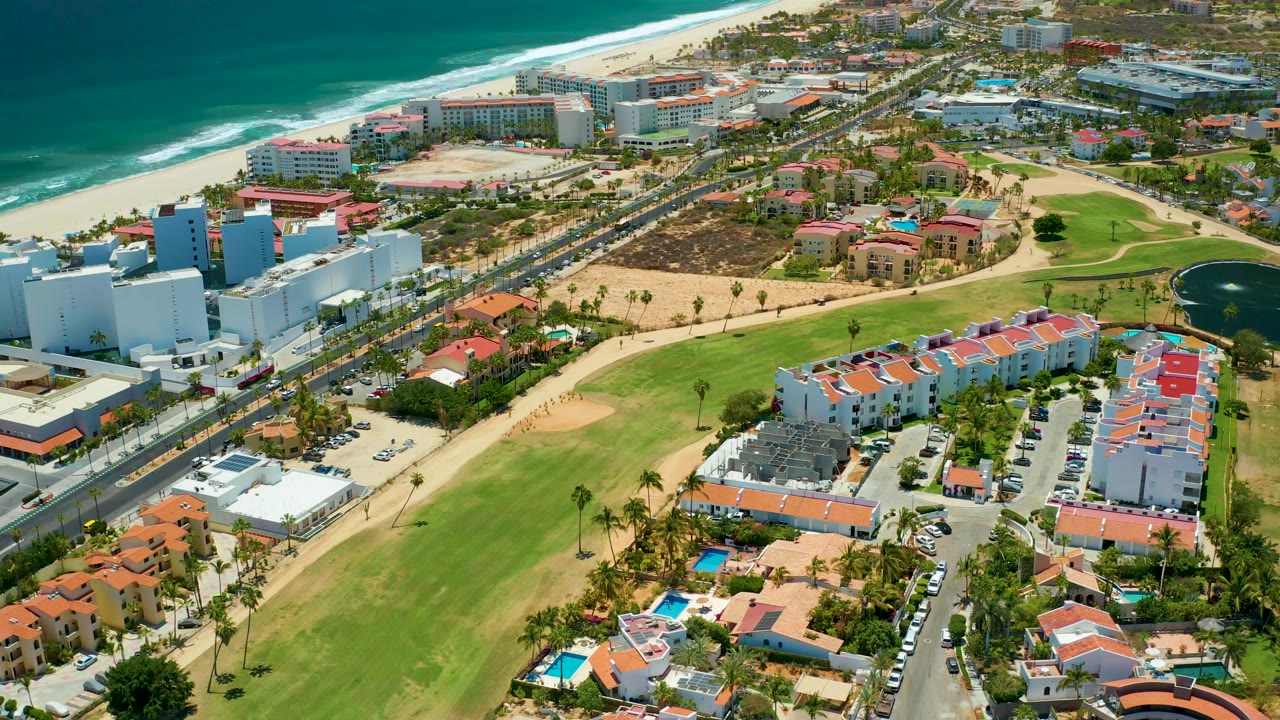 A slightly later aerial frame in San Jose Del Cabo, Mexico, keeps the surf line, sandy beach, road traffic, white resort-scale buildings, red-roof homes, lawns, palms, and pond visible, with no visible people.