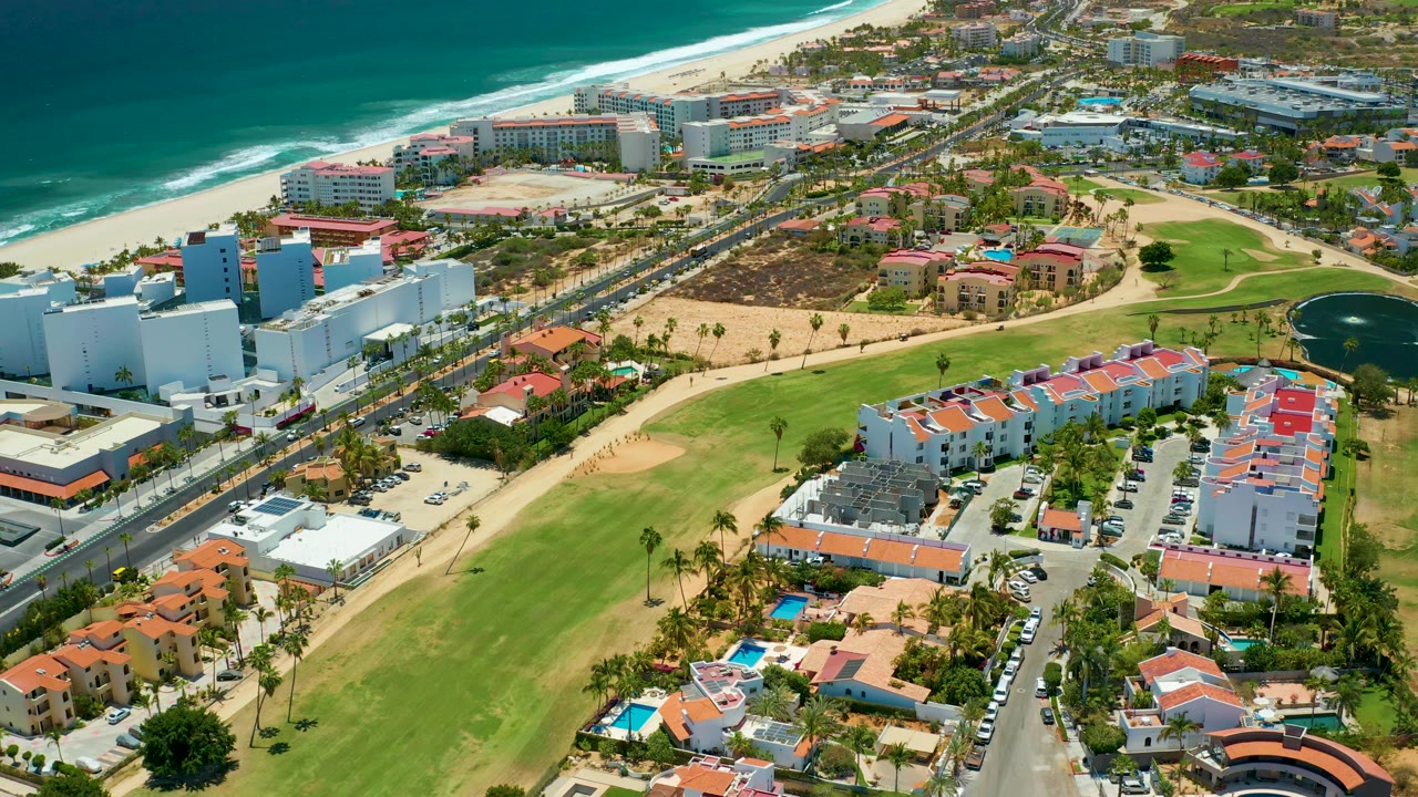 Aerial frame over San Jose Del Cabo, Mexico, with turquoise water and surf above the beach, a road crossing the middle, red and white buildings below, clipped grass areas, palm trees, and a small pond on the right, with no visible people.