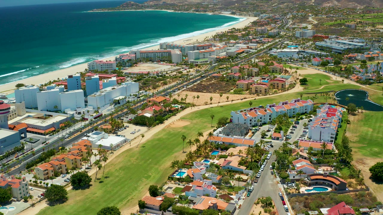 Opening aerial frame over San Jose Del Cabo shows blue ocean, white surf, sandy beach, coastal buildings, a road with vehicles, green lawns, red-roofed residences, distant hills, and no visible people.