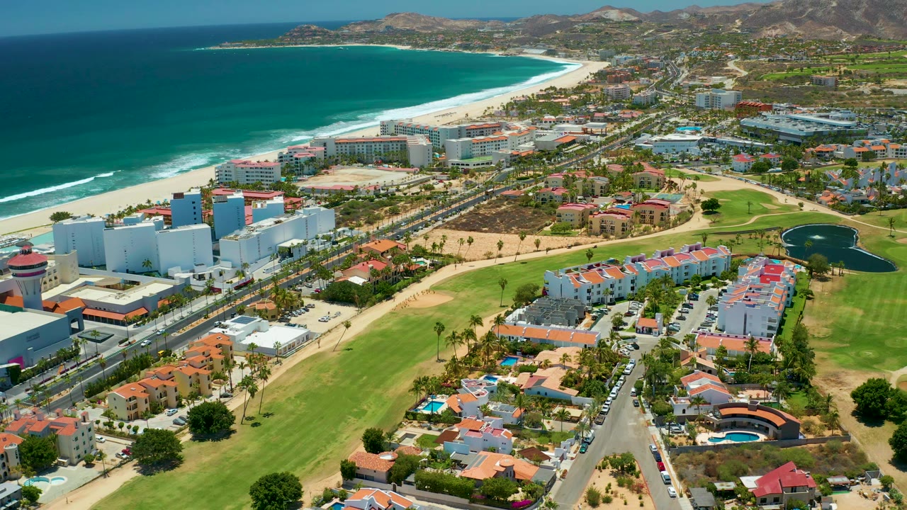 Coastal aerial scene with beach, ocean, roadside buildings, grass, trees, residences, and hills.