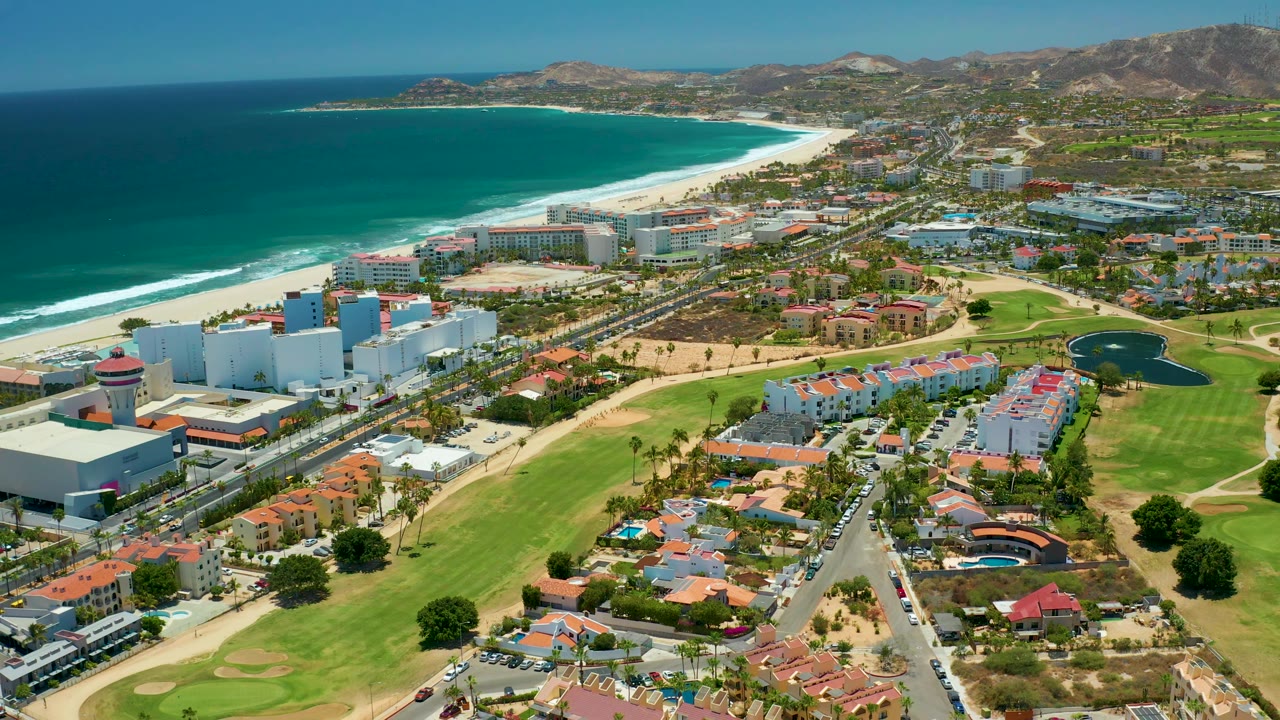 San Jose Del Cabo aerial view with the coastline stretching into the distance, a busy coastal road through the center, golf-course greens in the foreground, resort-style buildings, homes, hills, and no visible people.