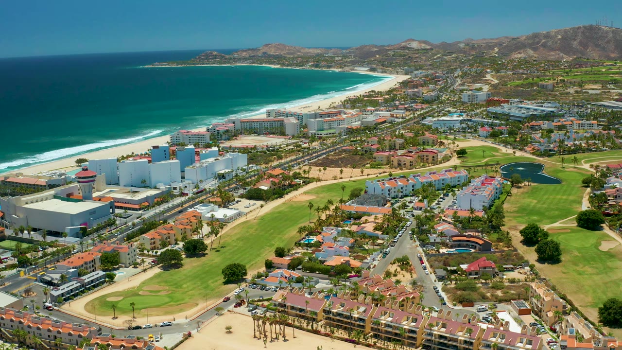 San Jose Del Cabo aerial frame with the beach and ocean stretching across the left side, white resort blocks below, green fairways and roads in the foreground, hills beyond town, and no visible people.