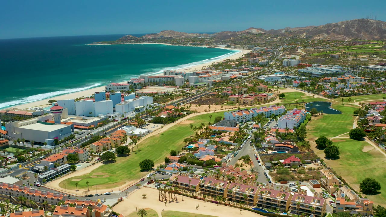 Aerial coastal town view with ocean, beach, resorts, roads, golf greens, and hills.