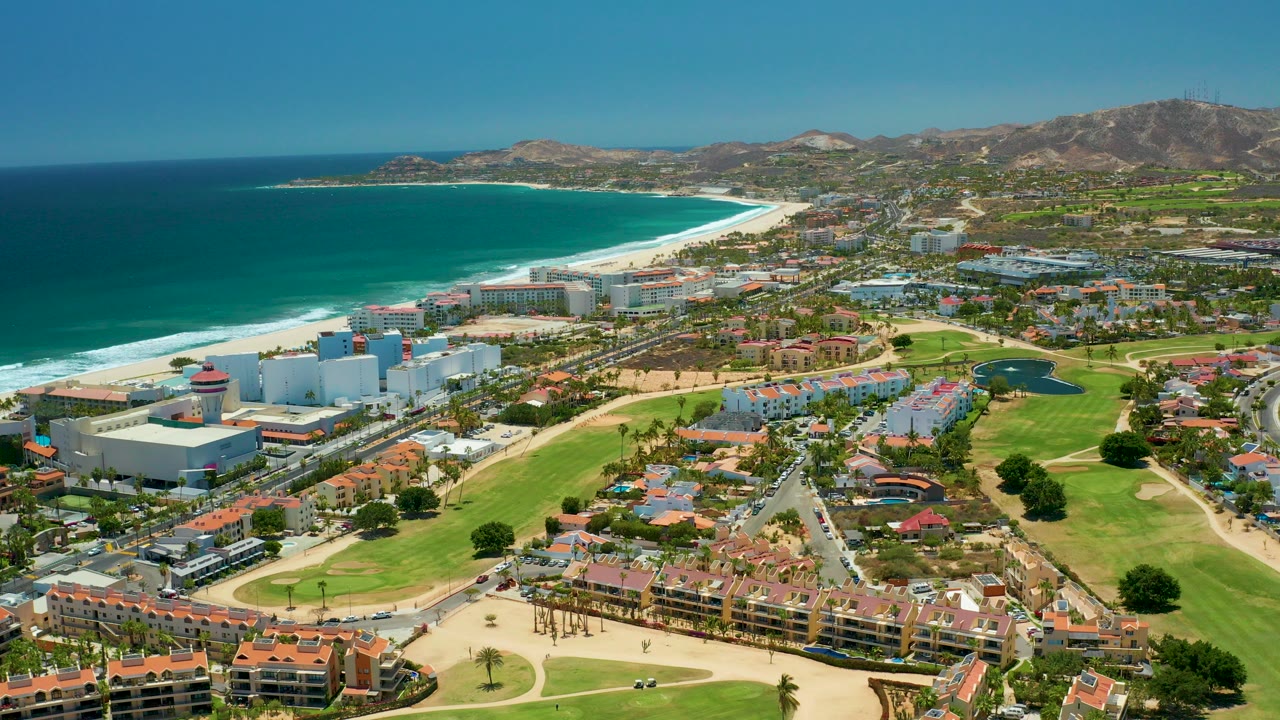 Opening aerial frame over San Jose del Cabo, Mexico, with turquoise ocean at left, a bright sandy beach, white resort buildings, green golf course fairways, red-roofed residences, distant hills, and no visible people.