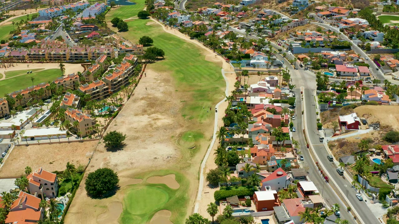 Aerial frame in San Jose Del Cabo with the golf course occupying the left side, a residential road on the right, and cars spaced along the pavement, with no visible people.