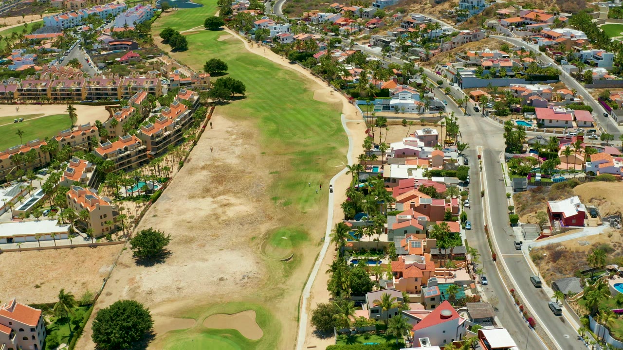 High aerial frame over San Jose del Cabo showing a road beside red-roofed homes, palms, green golf course grass, sandy patches, and no visible people.
