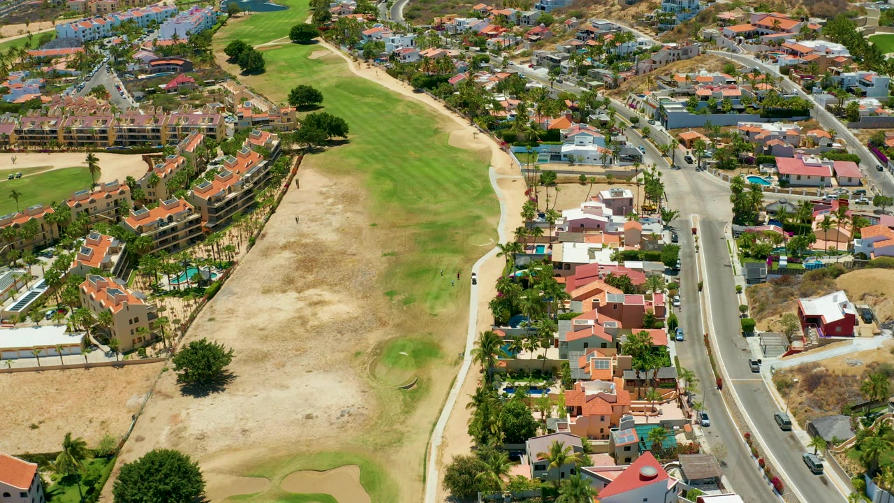 San Jose del Cabo aerial still with the road running down the right side, compact homes and palms beside it, golf course grass and sand on the left, and no visible people.