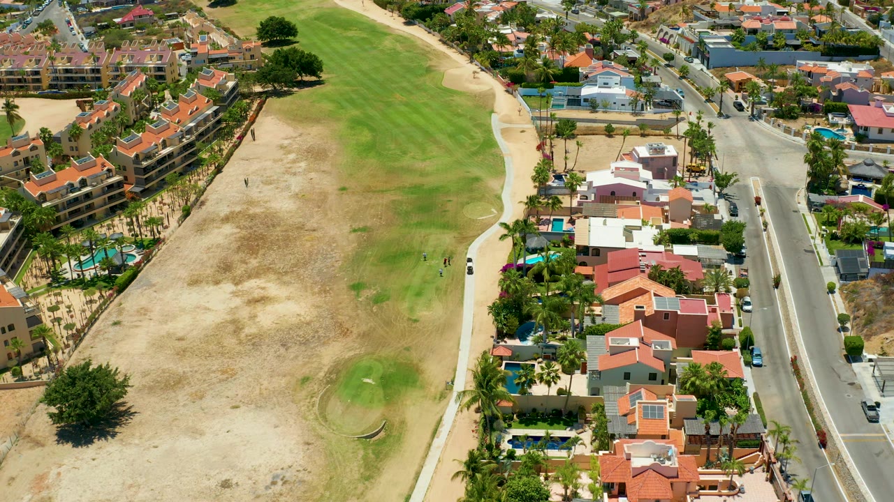 San Jose del Cabo aerial view with the pale walking path between a patchy golf course and a line of homes, while cars and other vehicles appear along the parallel road.