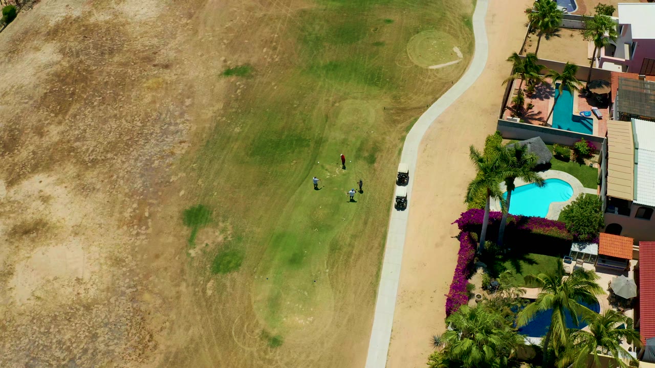 A high aerial frame in San Jose Del Cabo shows small people on a patchy grassy lot, a pale road, palm trees, private homes, and bright turquoise residential pools along the right edge.