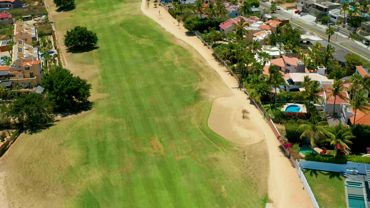 Aerial view one second in over the green fairway and right-side sand bunker in San Jose del Cabo, with homes, palms, a road, and no visible people.