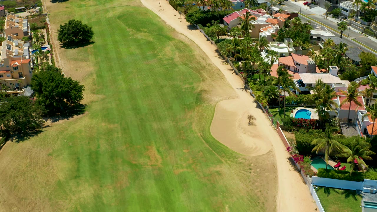 Mid-clip aerial frame shows more of the fairway center, the sand bunker near the right edge, San Jose del Cabo homes, pools, palms, and no visible people.