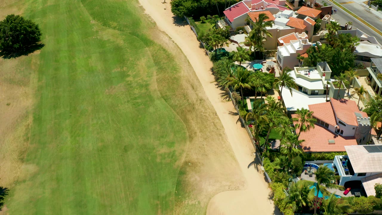 Overhead view in San Jose del Cabo showing a striped green field on the left, a sandy path, and white and red-roofed homes with palms on the right, with no visible people.