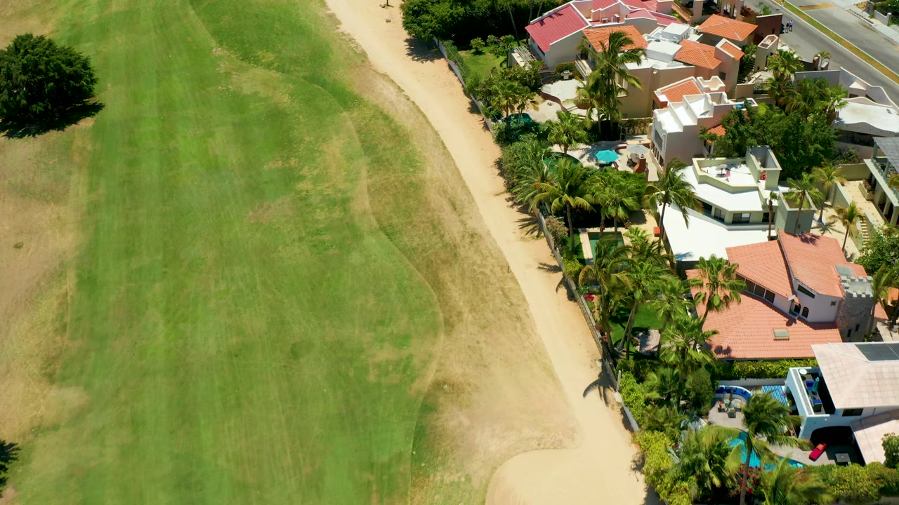 San Jose del Cabo aerial frame with the green field filling the left side, a pale sandy path through the center, and homes, palm trees, and a small blue pool on the right, with no visible people.