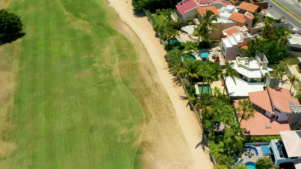 Overhead San Jose del Cabo view with the field's striped grass on the left, the sandy path running diagonally, and compact homes with palms and pools along the right edge, with no visible people.