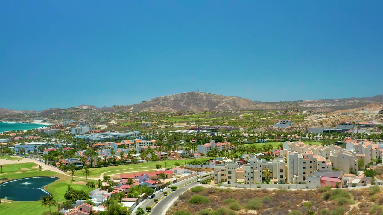 San Jose del Cabo aerial frame with white and tan buildings centered, golf fairways and ponds around them, ocean water on the left edge, and mountains across the background with no visible people.
