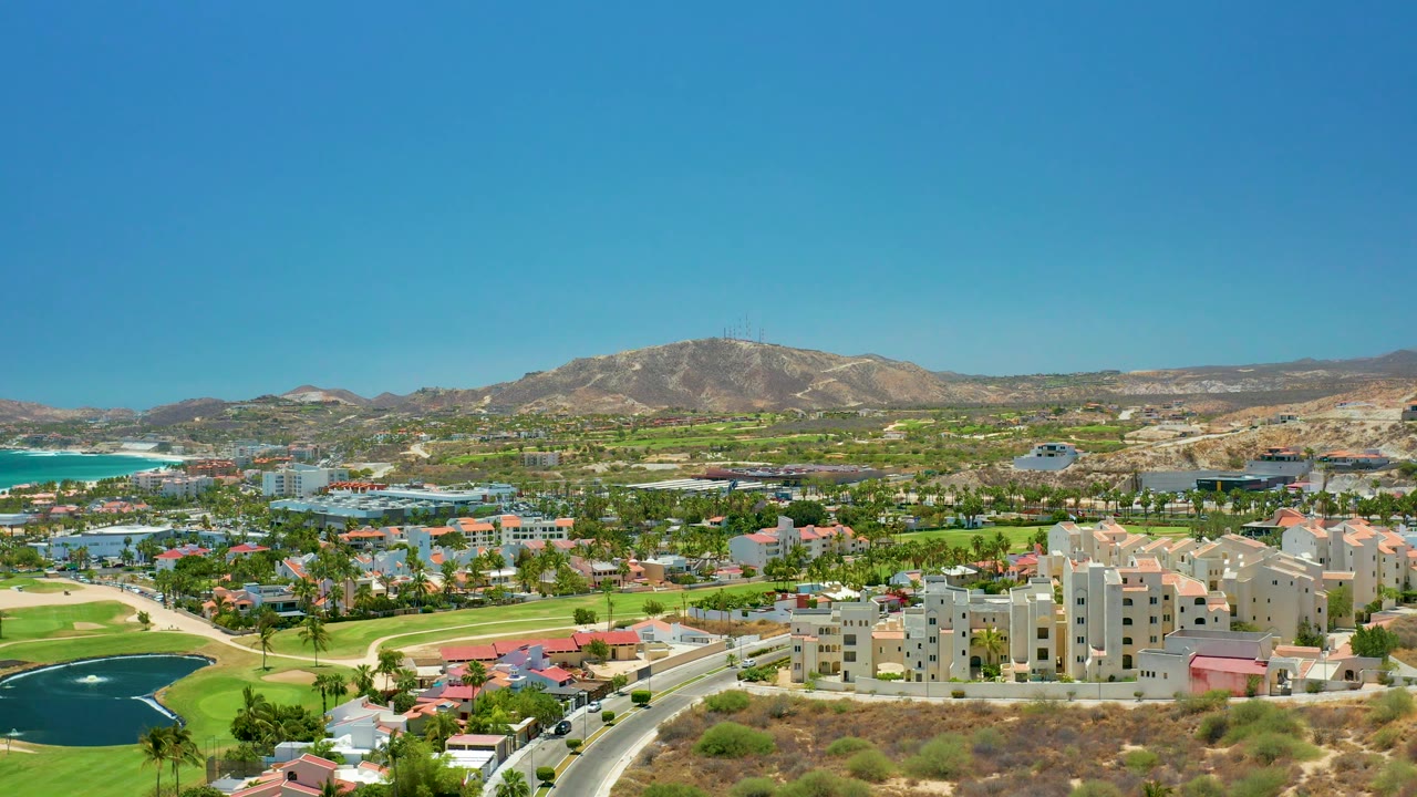 Wide San Jose del Cabo drone view with clustered buildings in the lower center, a curving road with small vehicles, green fairways, dark water hazards, turquoise ocean at left, and dry mountains beyond with no visible people.