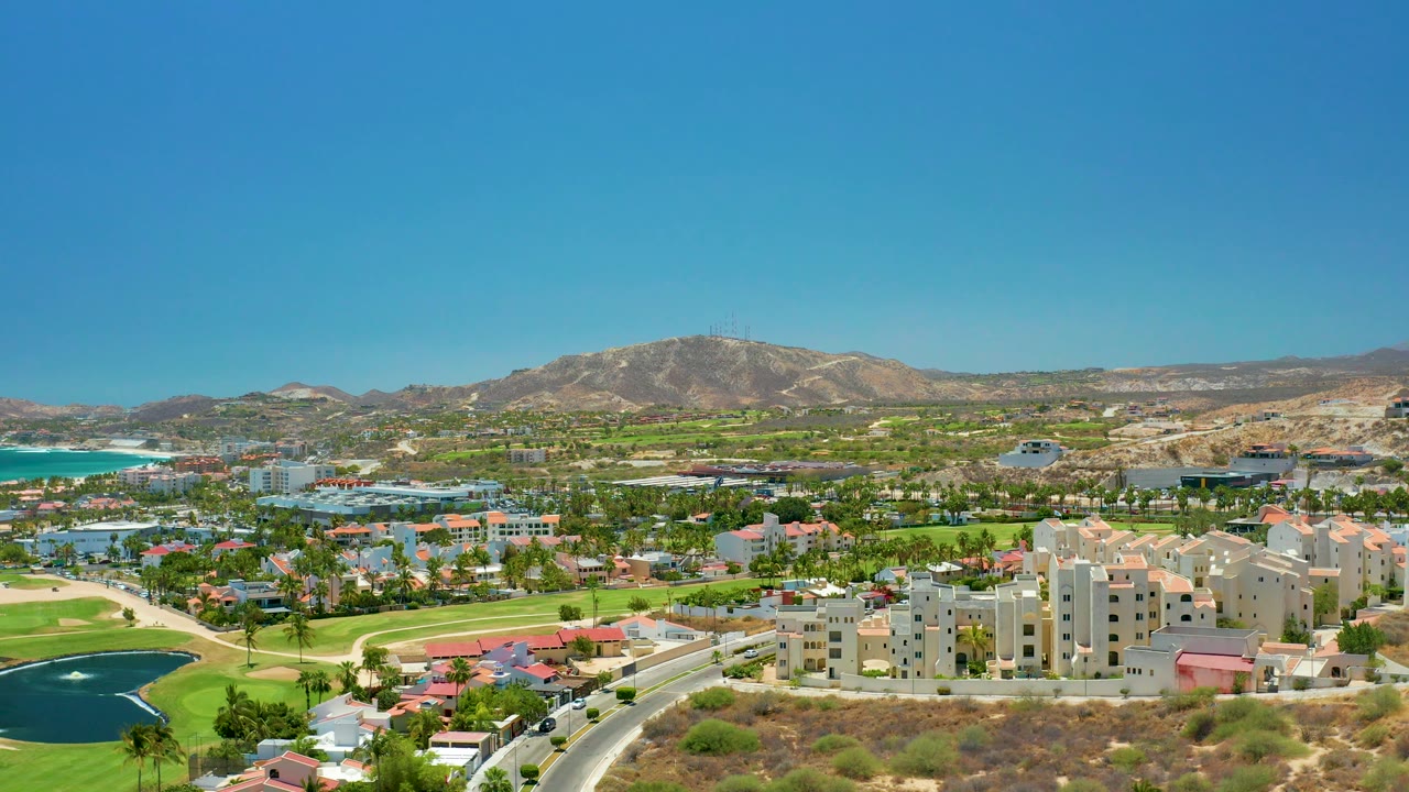 Aerial landscape with golf fairways, coastal water, buildings, road, and mountain backdrop.