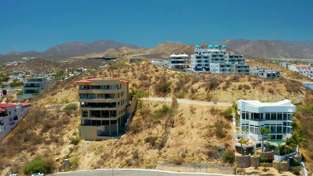 Opening aerial frame over San Jose del Cabo hillside residences, with tan and white modern buildings on dry slopes, distant mountains, blue sky, and no visible people.