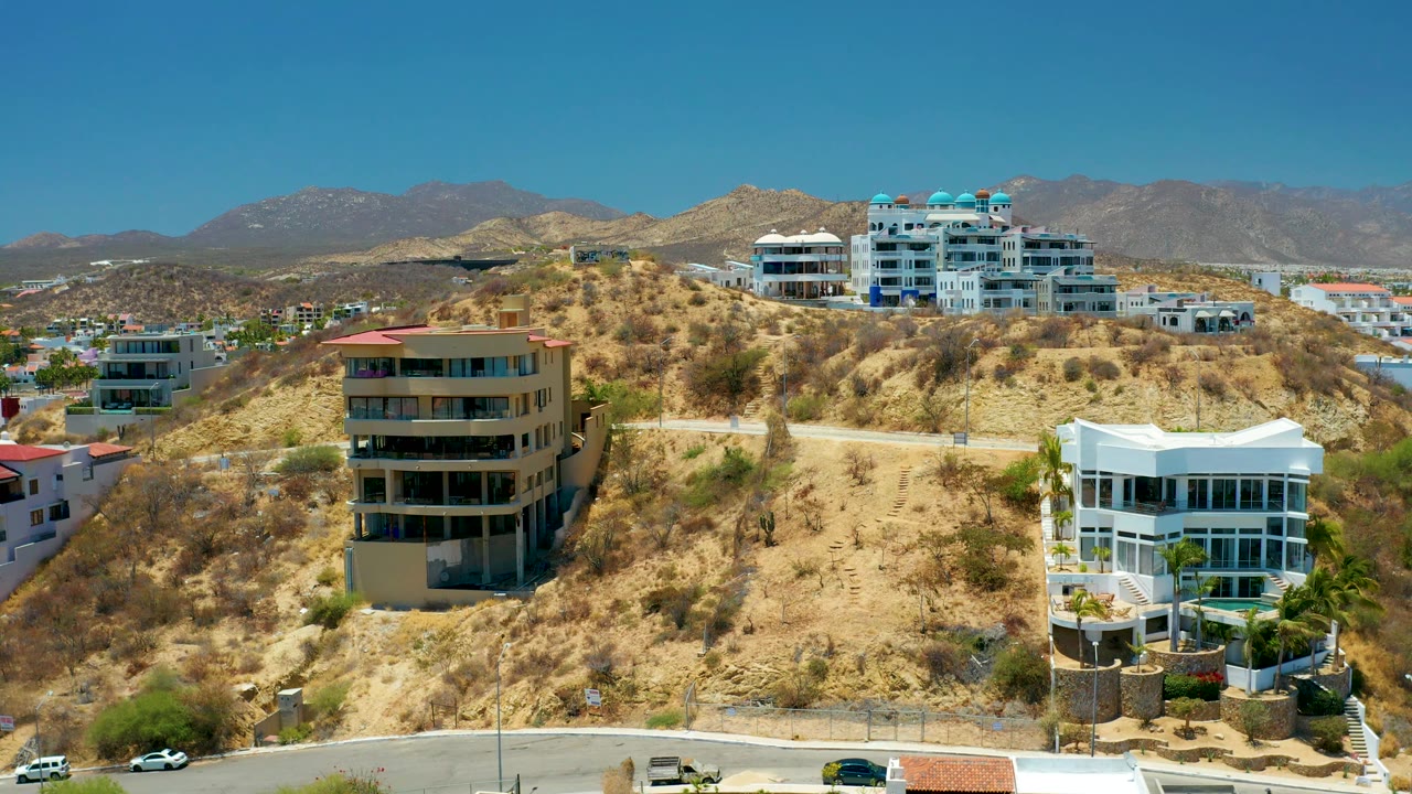 San Jose del Cabo aerial frame showing hillside homes spread across arid brown ground, a road at the base, mountain ridges in back, and no visible people.