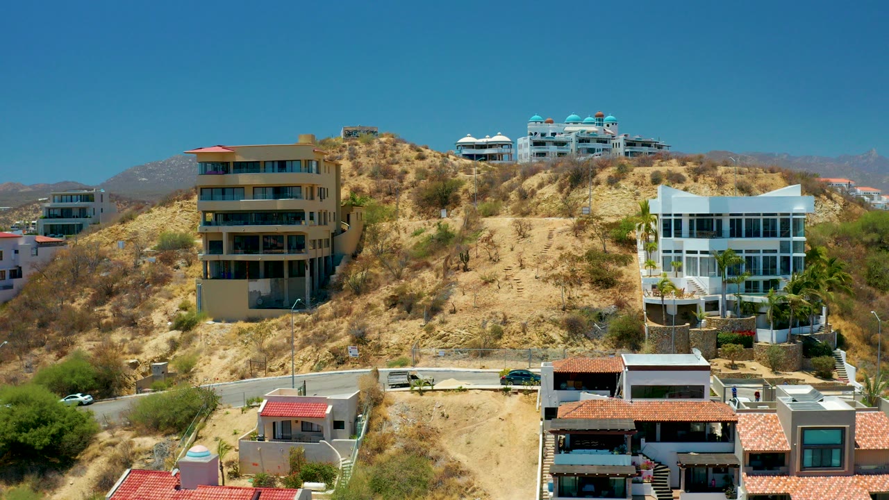 Opening frame of modern hillside residences in San Jose Del Cabo, with a tan slope, red roof sections, white buildings, distant hills, and no visible people.