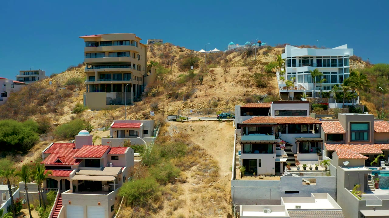 San Jose Del Cabo residential buildings sit across a tan hillside with a dirt road near the upper slope, red roofs below, and no visible people.