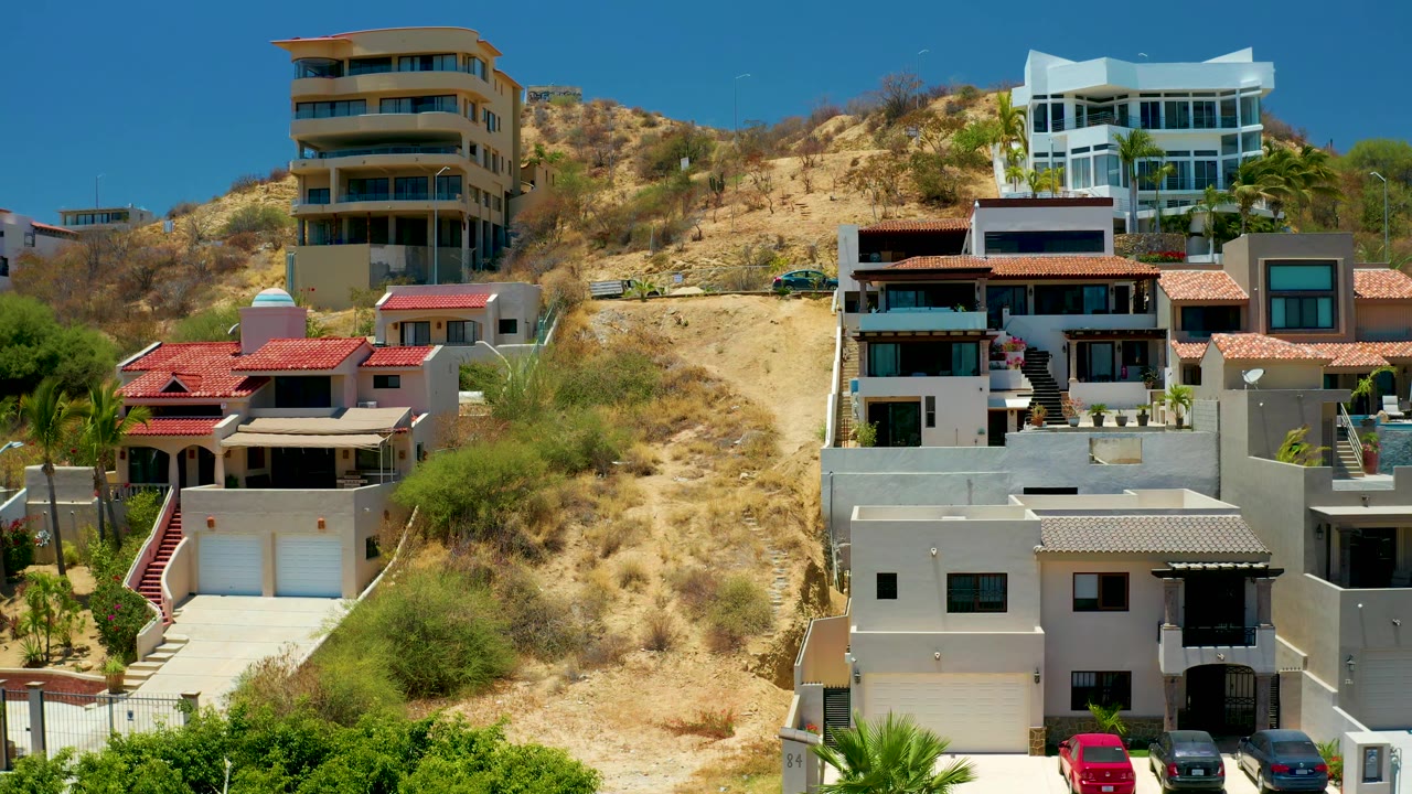 San Jose Del Cabo hillside housing with tan and white buildings, balconies, a central dry slope, green plants along the lower edge, and no visible people.