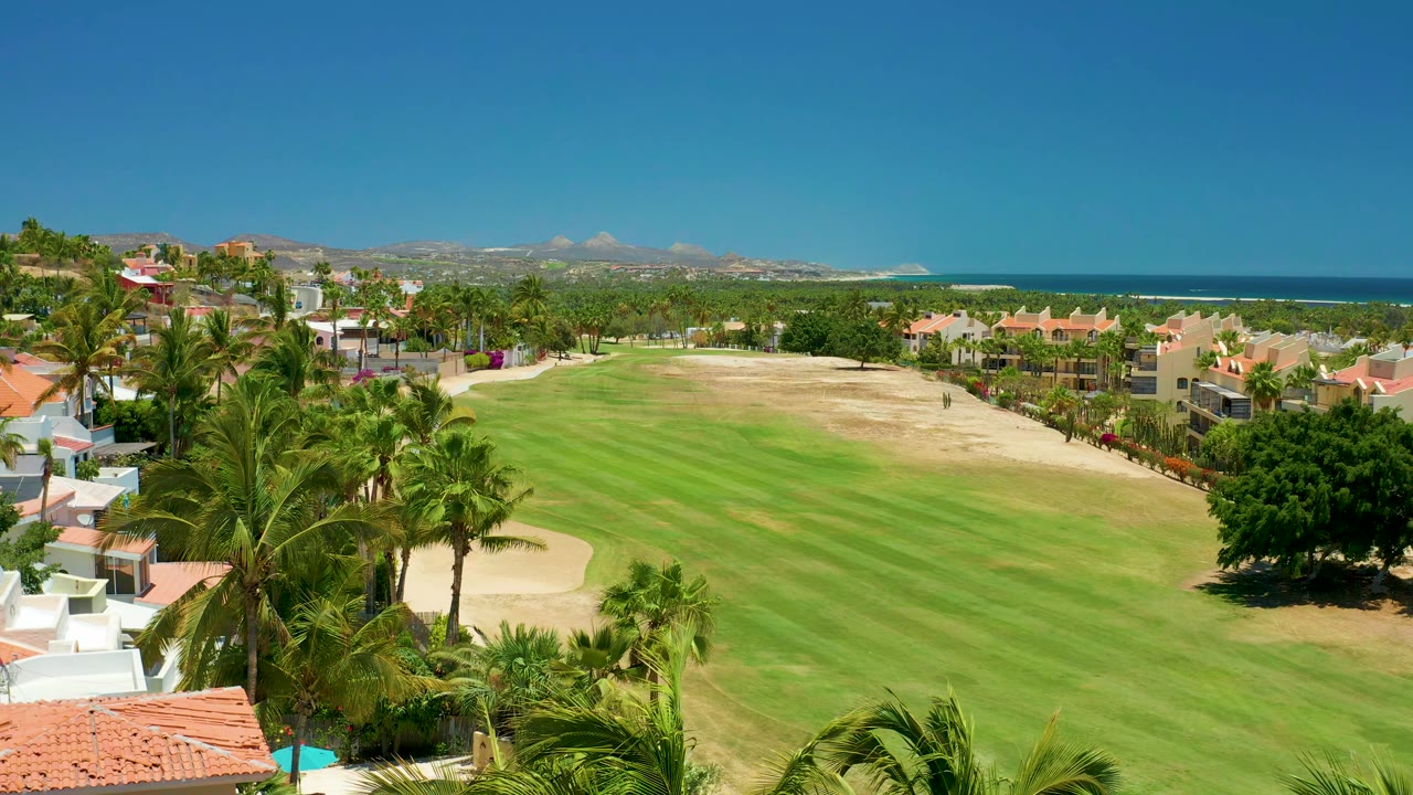 Golf fairway bordered by palms and red-roofed buildings under a clear blue sky.