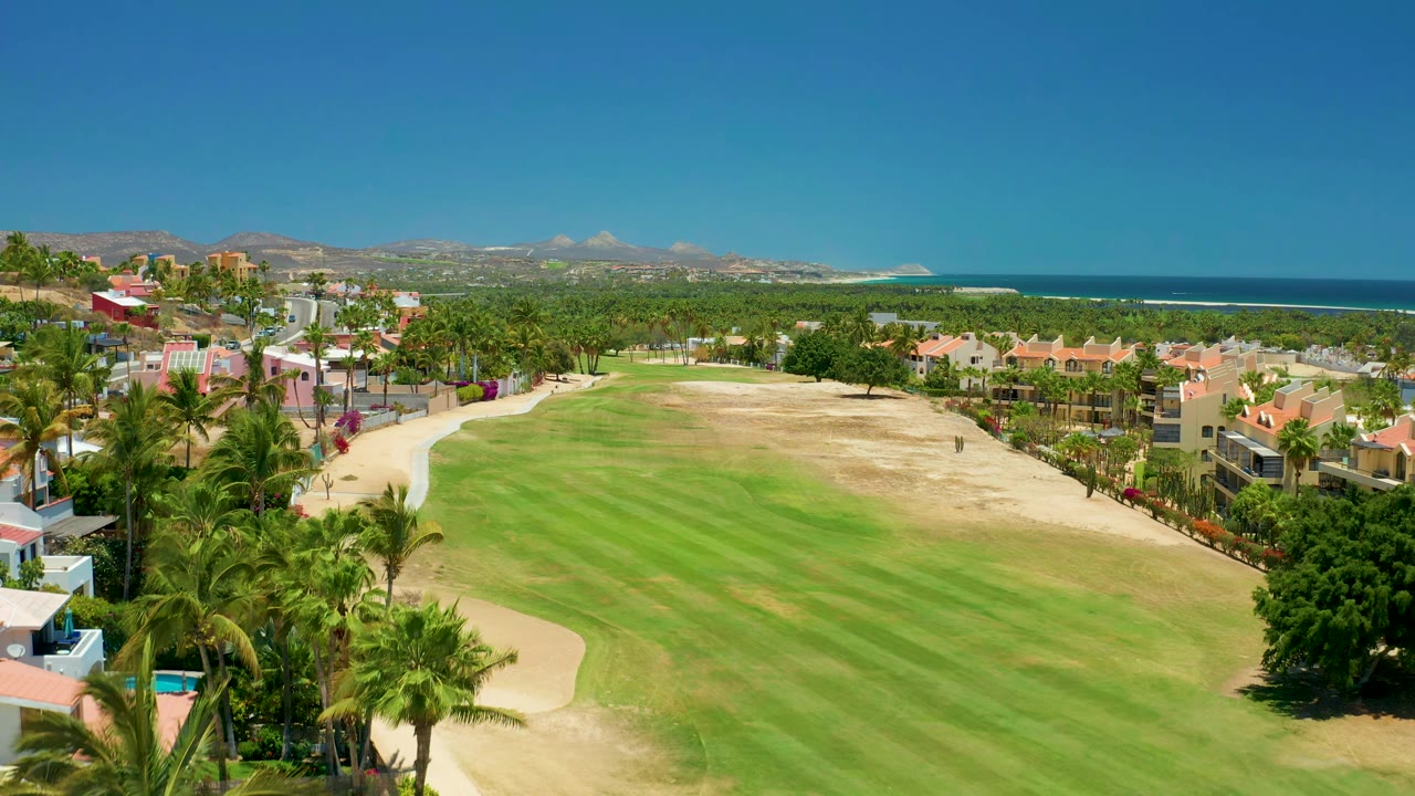 Wide aerial view over a golf course, palm-lined homes, ocean, and hills.