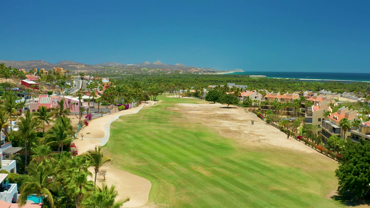 A San Jose del Cabo aerial angle shows the dry fairway shifted right, palm trees and residential buildings in the foreground, ocean along the horizon, and no visible people.