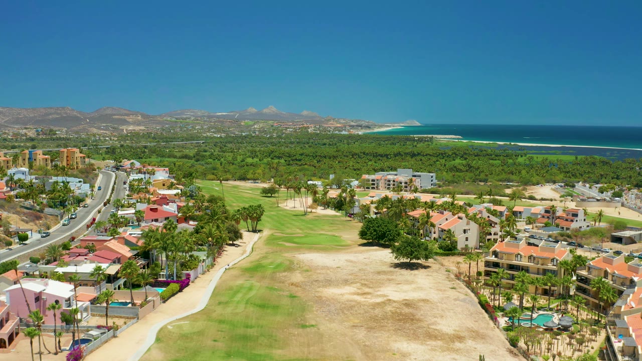 San Jose Del Cabo aerial view with the road slightly more central, small vehicles on pavement, houses around the golf course, distant mountains, ocean, and no visible people.