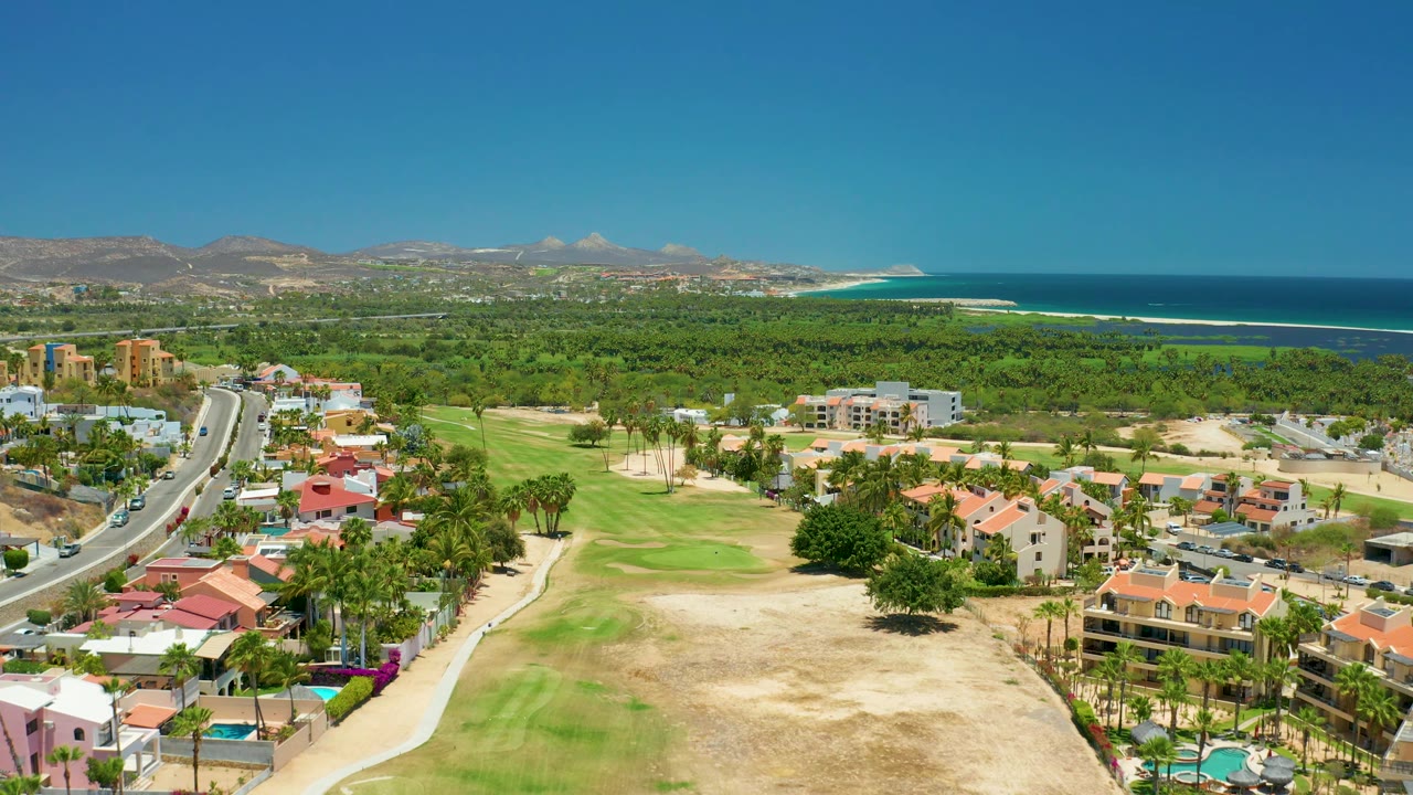 Aerial frame over San Jose Del Cabo showing red-roof housing in the lower frame, a road with cars on the left, golf course greens, wooded land, mountains, ocean, and no visible people.