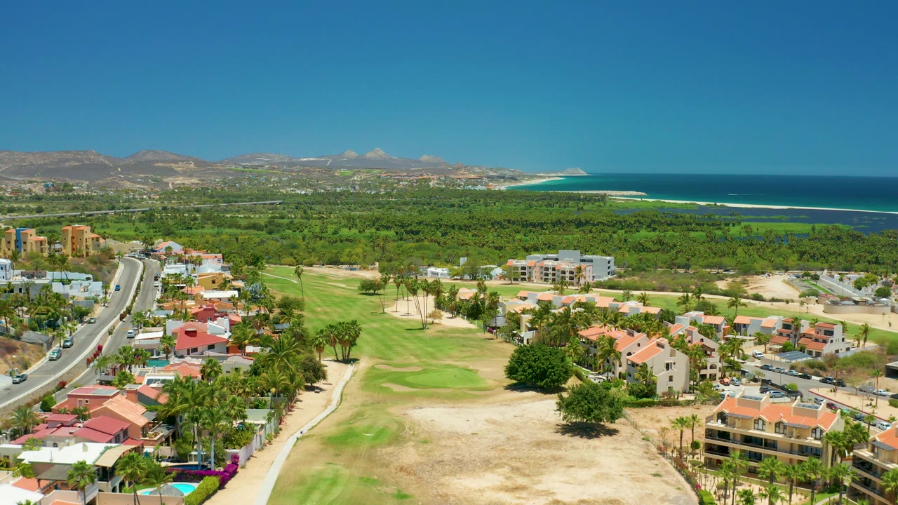 Aerial view across homes, road, golf course, dense trees, mountains, and blue ocean.