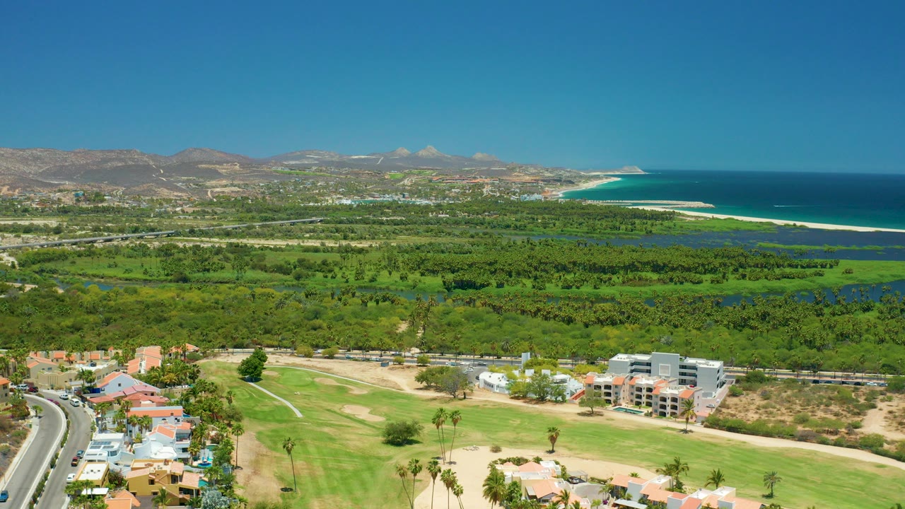 Opening aerial frame over San Jose Del Cabo showing foreground homes and golf fairways, dense green vegetation, distant mountains, coastline, blue ocean, and no visible people.