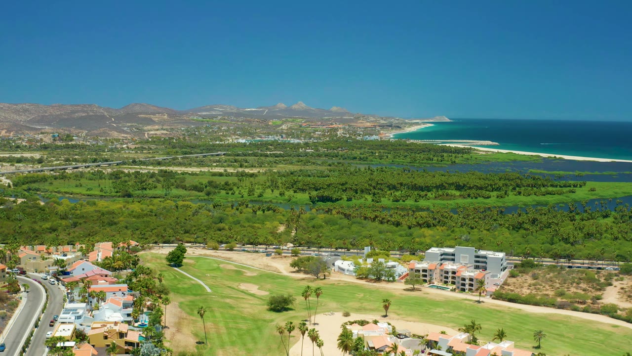 Drone view across golf greens, residential buildings, vegetation, mountains, and ocean.