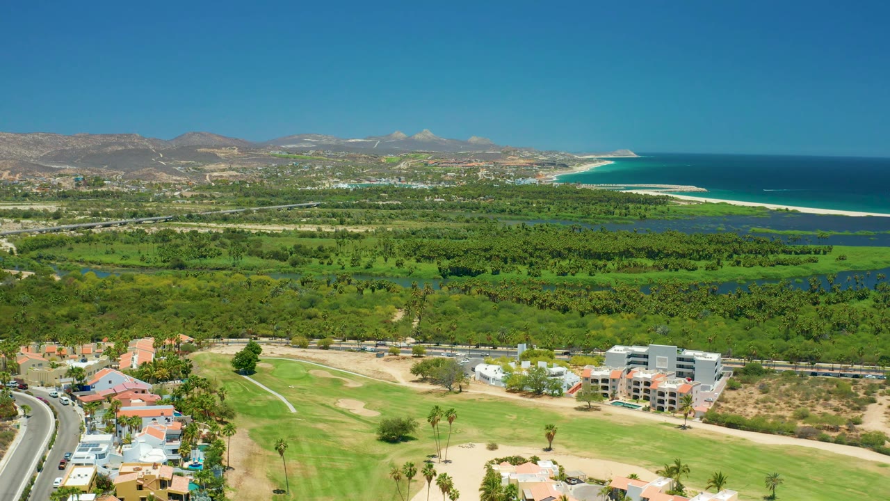 San Jose Del Cabo aerial view with residential roofs and a golf course in the lower frame, broad green vegetation and water channels beyond, mountains in back, ocean to the right, and no visible people.