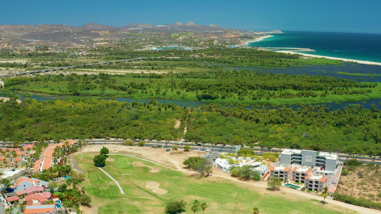 Aerial view over San Jose Del Cabo with a golf course and buildings in the foreground, green waterway vegetation through the center, beach and ocean at right, hills beyond, and no visible people.