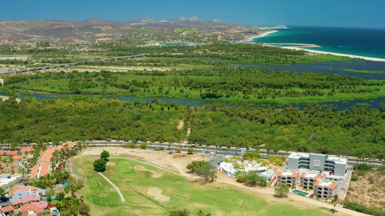 Aerial view in San Jose Del Cabo with the coastline more prominent on the right, green waterways across the center, golf course and multi-story buildings at the bottom, distant hills, and no visible people.