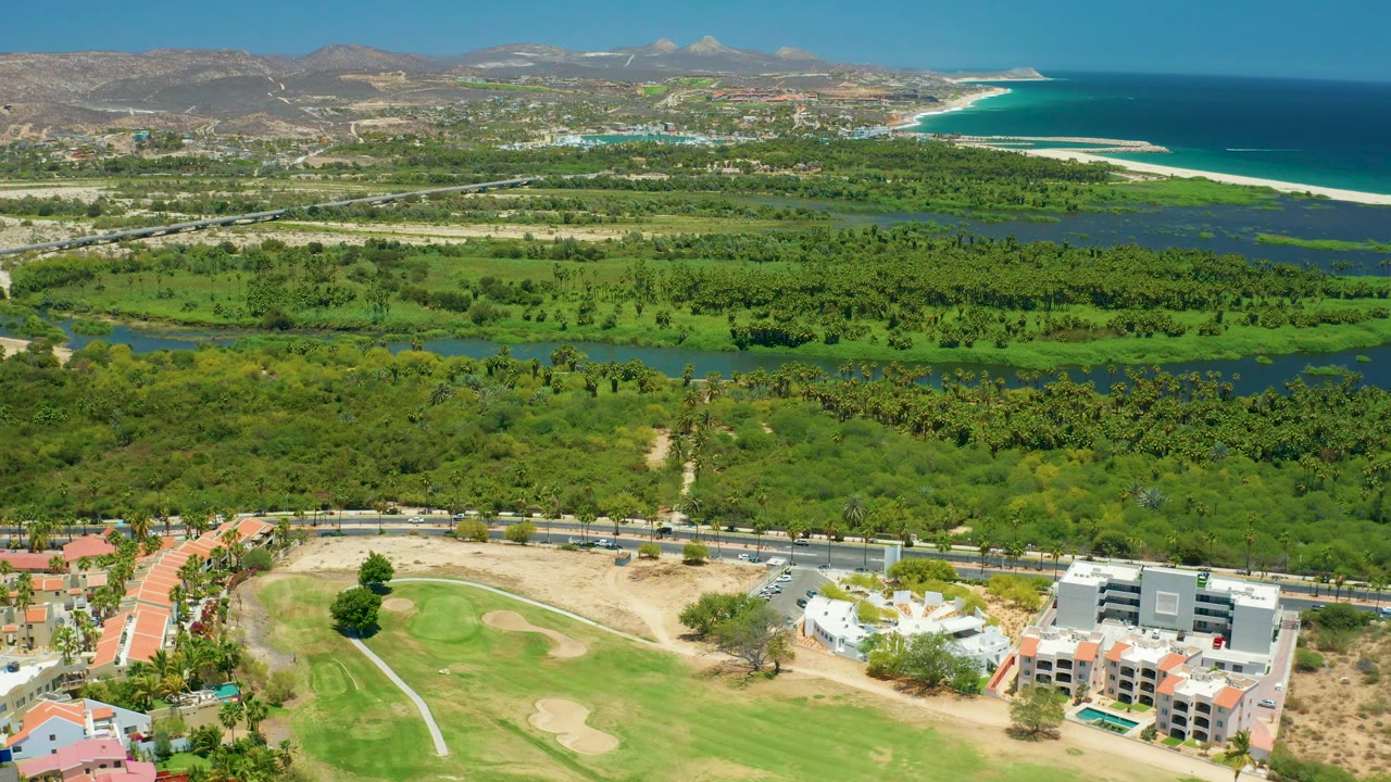 Drone landscape with golf green, buildings, road, dense vegetation, waterway, beach, and ocean.