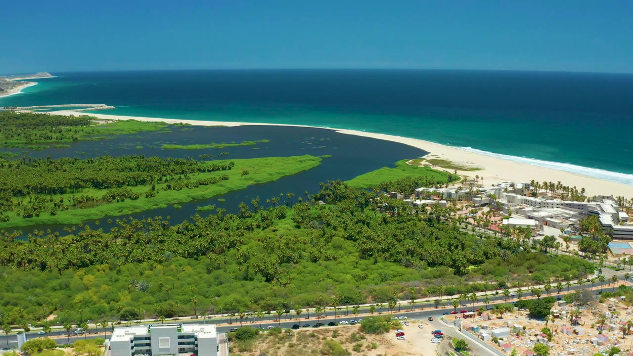 Wide aerial frame of San Jose Del Cabo with green vegetation around a dark lagoon, a bright sand beach, turquoise ocean, road traffic, low buildings, and no visible people.